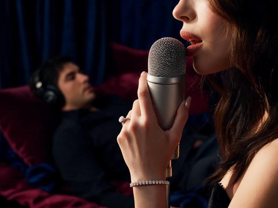 Composição cinematográfica mostrando uma mulher elegante em ambiente luxuoso, segurando um microfone profissional próximo aos lábios de forma sedutora. Ao fundo, silhueta masculina deitada na cama usando fones de ouvido, transmitindo a conexão íntima através do áudio. Iluminação dramática em tons de