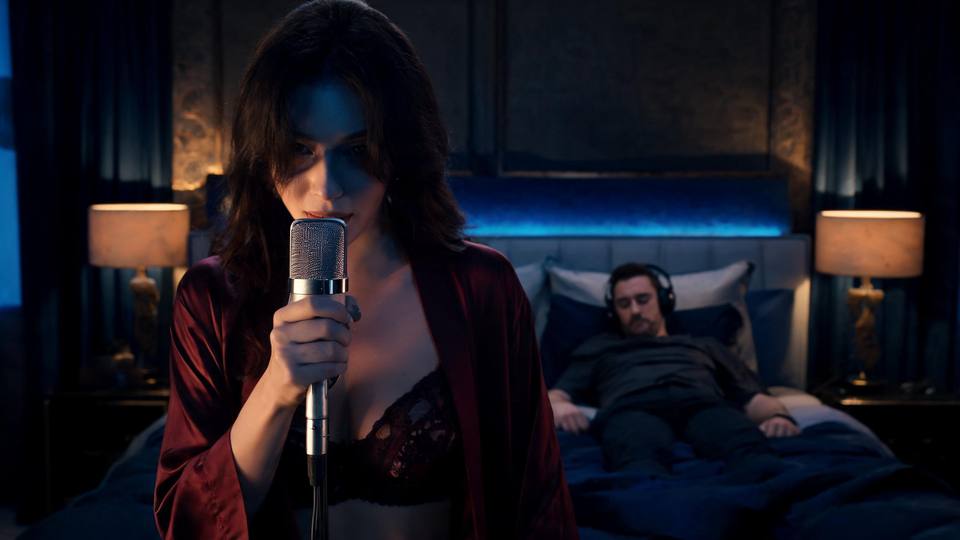 Close elegante mostrando lábios femininos próximos ao microfone em pose sensual de sussurro, com mão delicada segurando o equipamento. Ao fundo desfocado, silhueta de homem deitado usando fones de ouvido. A imagem transmite sedução através da voz e intimidade discreta.