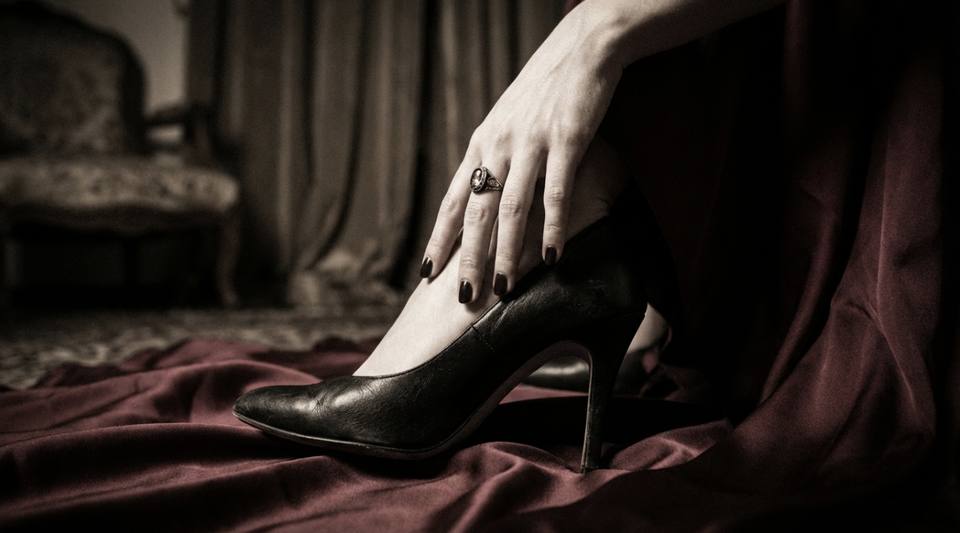 Close-up da mão de uma mulher apoiada no tornozelo enquanto usa salto alto preto sobre um tecido vermelho escuro.